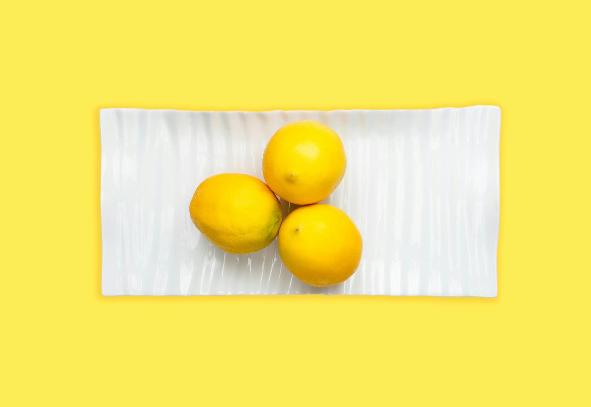 Tres limones frescos sobre un plato blanco con fondo amarillo vibrante, enfatizando frescura y cítricos.