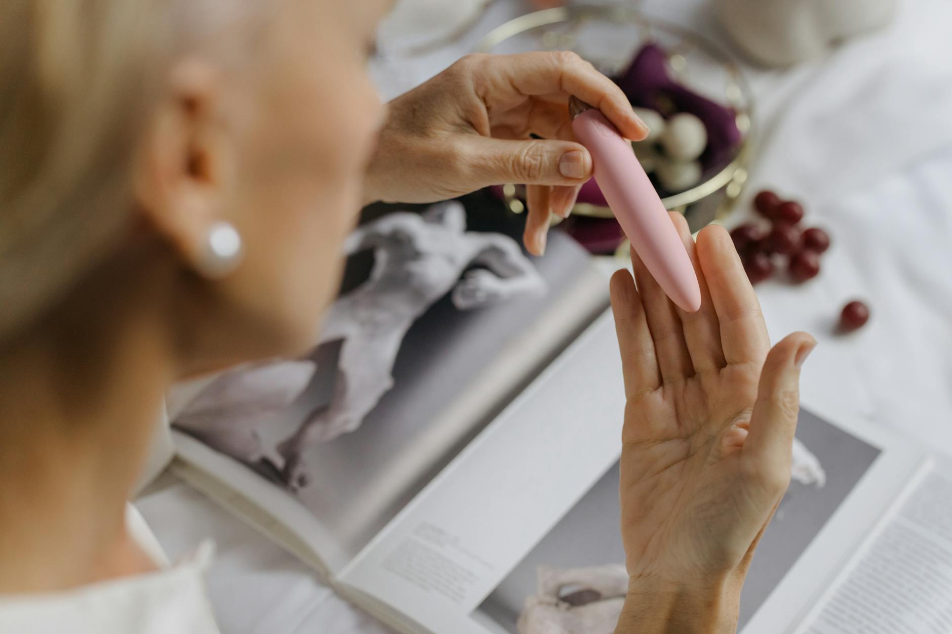 Detalle de una mujer sosteniendo un vibrador rosa, con libros de arte de fondo, enfatizando temas íntimos y personales