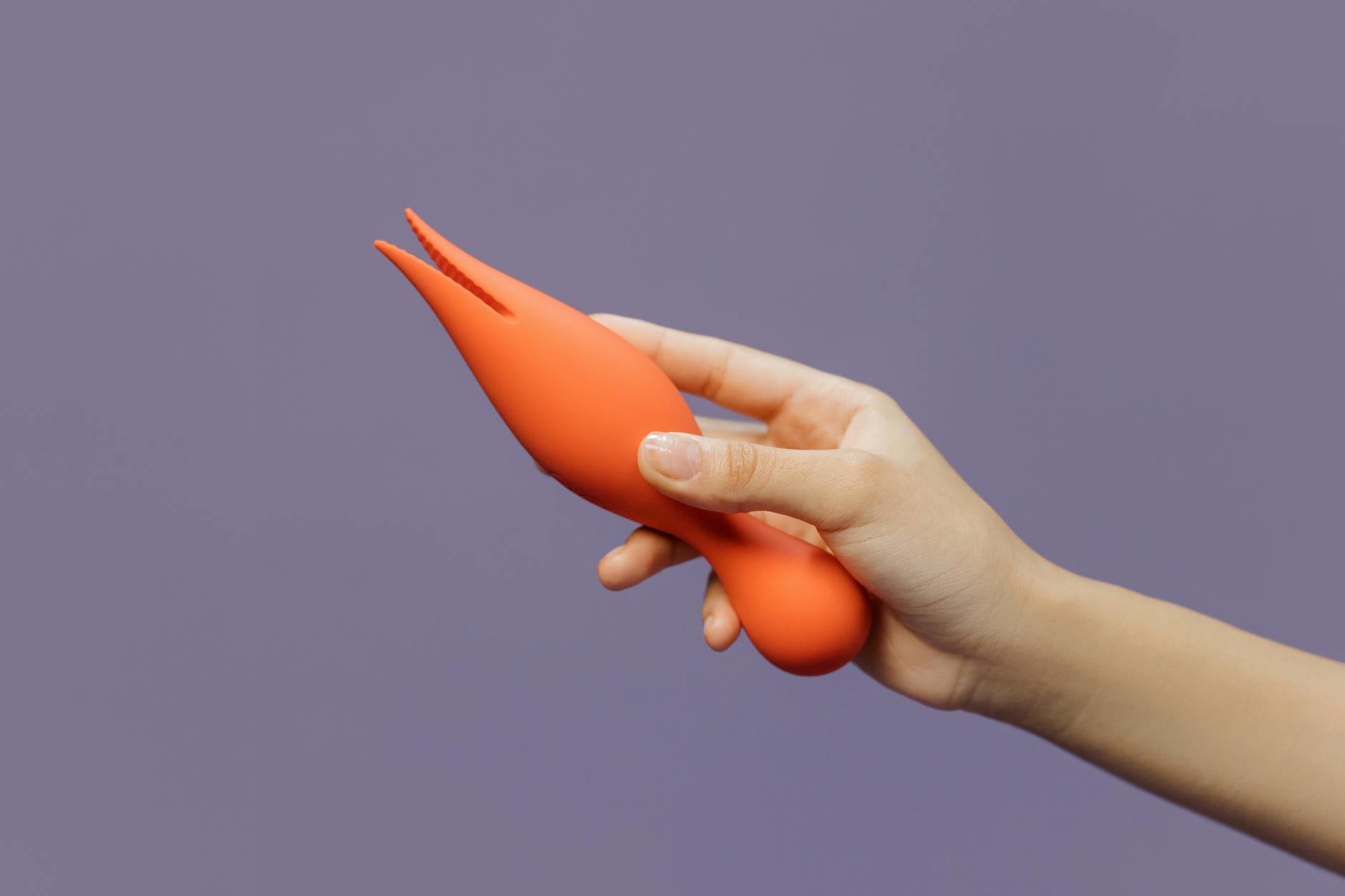 Mano sosteniendo un vibrador naranja contra un fondo minimalista violeta, mostrando sensualidad moderna