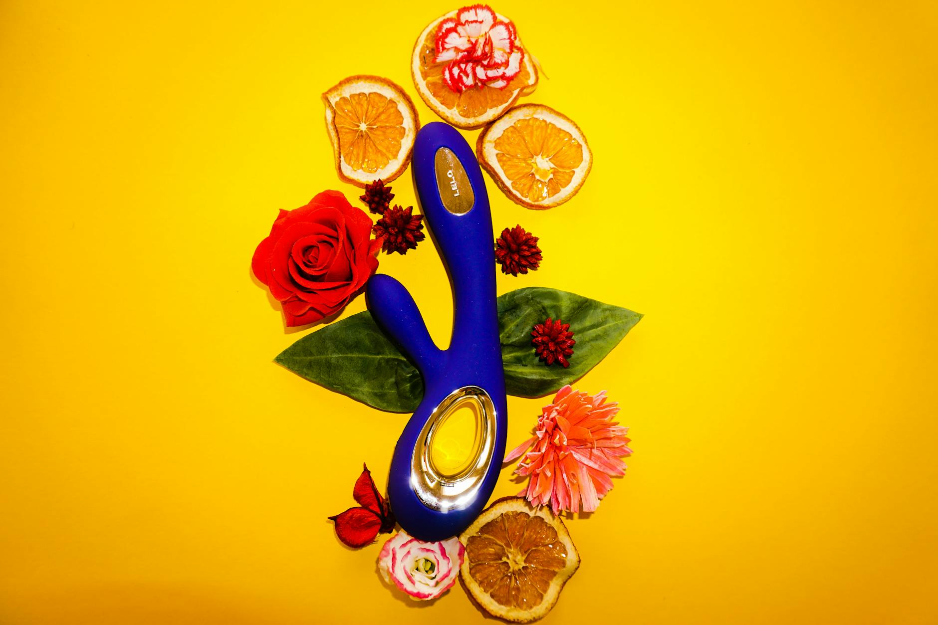 Foto de un vibrador azul rodeado de flores y naranjas sobre un fondo amarillo